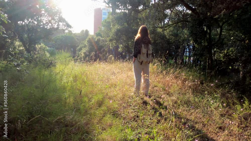 Hiking. A girl with a backpack stands on a forest path and enjoys a beautiful sunset. Golden light in the green forest. Forest hike. Sunny summer day. Walking among the trees and flowers. Holidays. Hiking. A girl with a backpack stands on a forest path and enjoys a beautiful sunset. Golden light in the green forest. Forest hike. Sunny summer day. Walking among the trees and flowers. Holidays.