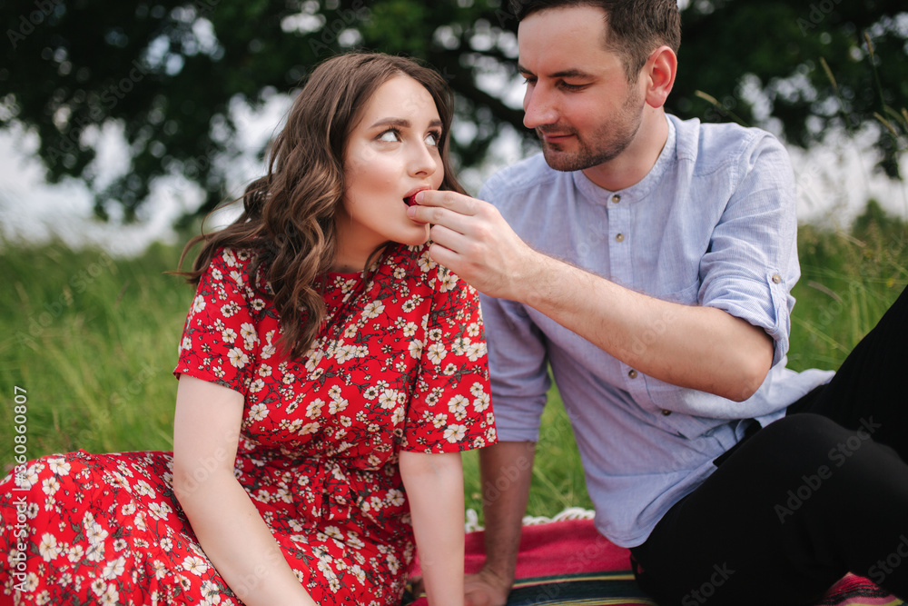 Attactive young woman with handsome man sits outdoors on red blanket. Woman taske strawbeerry Attactive young woman with handsome man sits outdoors on red blanket. Woman taske strawbeerry