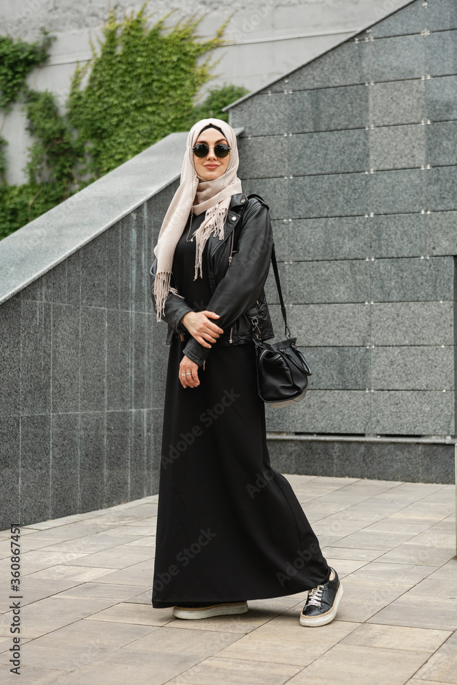 modern stylish muslim woman in hijab, leather jacket and black abaya walking in city street , wearing sunglasses and bag, fashion style trend - obrazy, fototapety, plakaty 