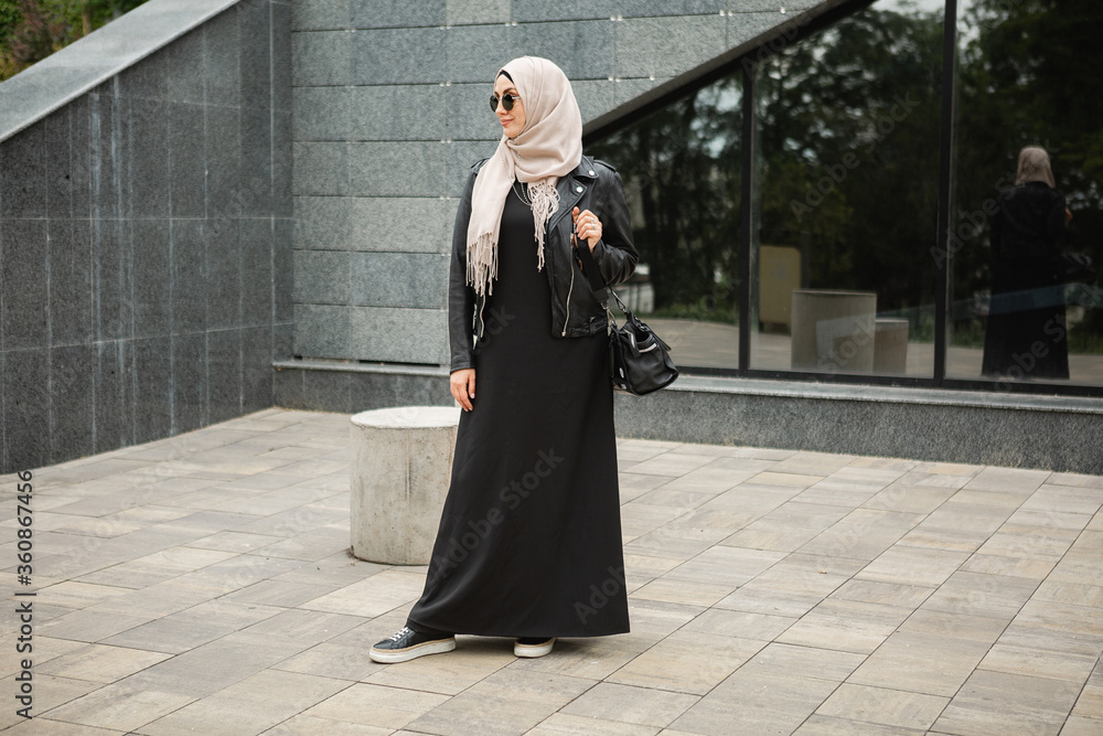 modern stylish muslim woman in hijab, leather jacket and black abaya walking  in city street , wearing sunglasses and bag, fashion style trend Stock  Photo | Adobe Stock