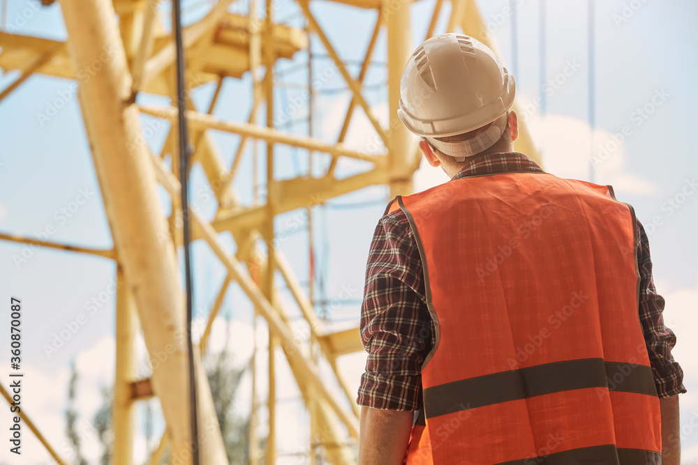 a construction engineer in an orange vest and hard hat stands with his back to the camera against the background of a yellow construction tower crane