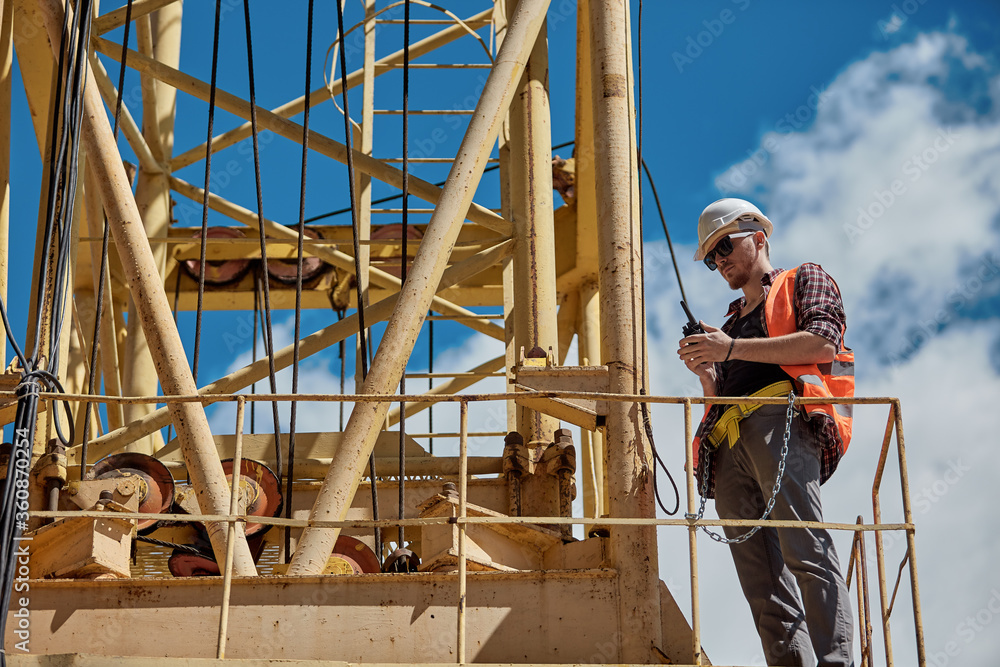 A construction engineer in a checked shirt, orange vest, and white hard hat on a yellow construction crane with a walkie-talkie in his hands. foreman, supervisor, worker