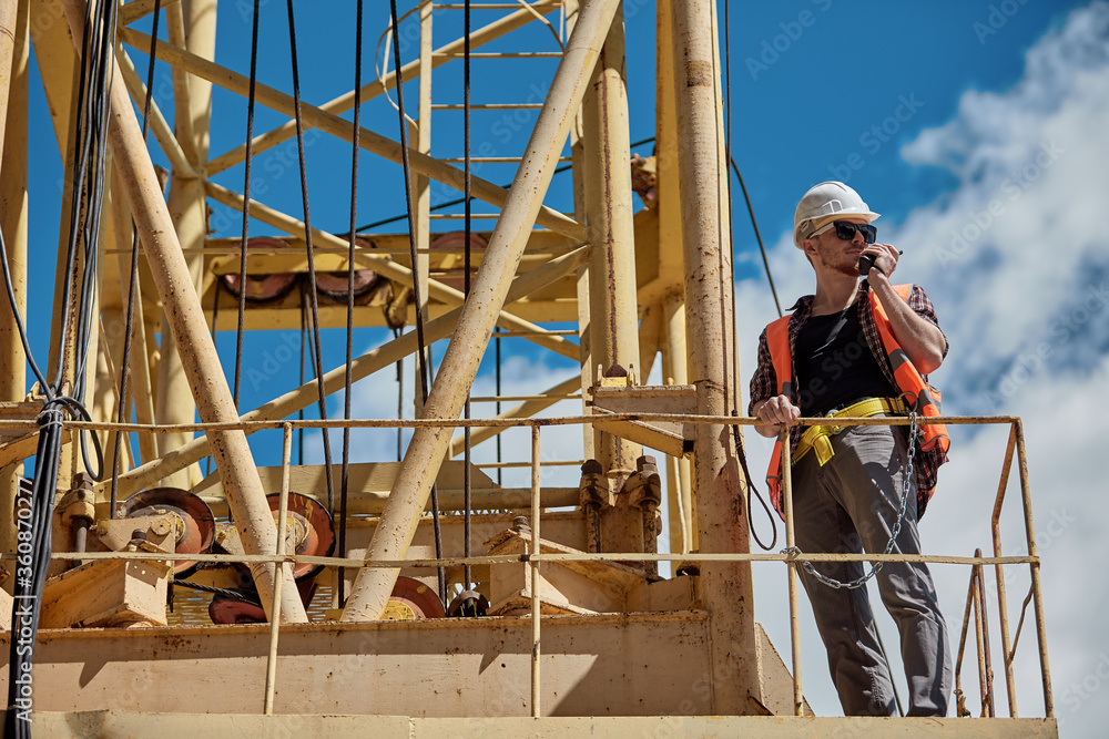 A construction engineer in a checked shirt, orange vest, and white hard hat on a yellow construction crane with a walkie-talkie in his hands. foreman, supervisor, worker A construction engineer in a checked shirt, orange vest, and white hard hat on a yellow construction crane with a walkie-talkie in his hands. foreman, supervisor, worker