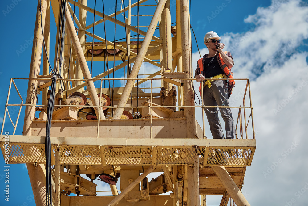 A construction engineer in a checked shirt, orange vest, and white hard hat on a yellow construction crane with a walkie-talkie in his hands. foreman, supervisor, worker A construction engineer in a checked shirt, orange vest, and white hard hat on a yellow construction crane with a walkie-talkie in his hands. foreman, supervisor, worker