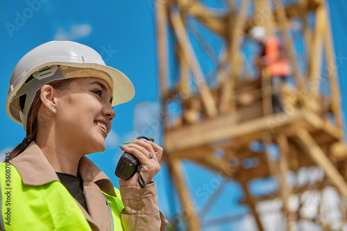 Close - up of a girl engineer with a walkie-talkie in hand on a construction site in a protective vest and hard hat, in the background a worker on a yellow crane