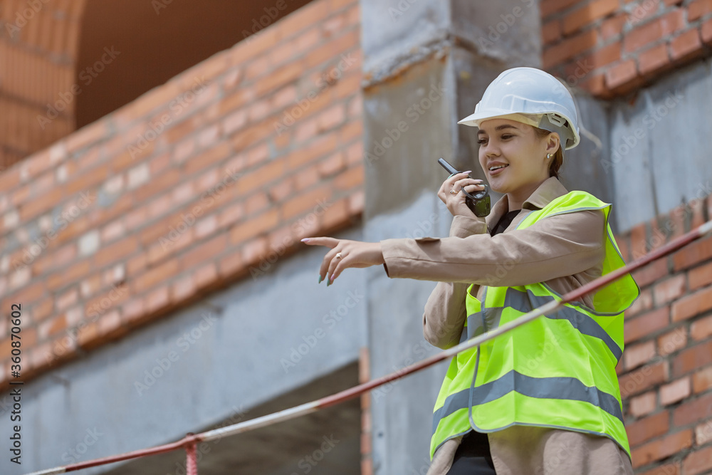 The girl is an engineer and a walkie talkie in hand on the construction site in a protective vest gives instructions pointing with her hand The girl is an engineer and a walkie talkie in hand on the construction site in a protective vest gives instructions pointing with her hand