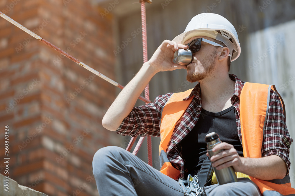 A civil engineer in a checked shirt, orange vest, and white hard hat. Happy young engineer, architect, worker sitting on a construction site and resting at lunch A civil engineer in a checked shirt, orange vest, and white hard hat. Happy young engineer, architect, worker sitting on a construction site and resting at lunch
