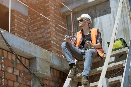 A civil engineer in a checked shirt, orange vest, and white hard hat. Happy young engineer, architect, worker sitting on a construction site and resting at lunch
