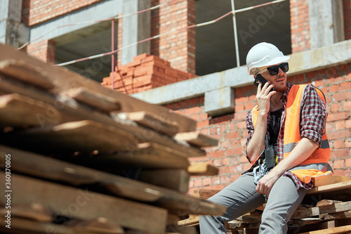 An engineer in a construction uniform talks on the phone against the background of a building under construction