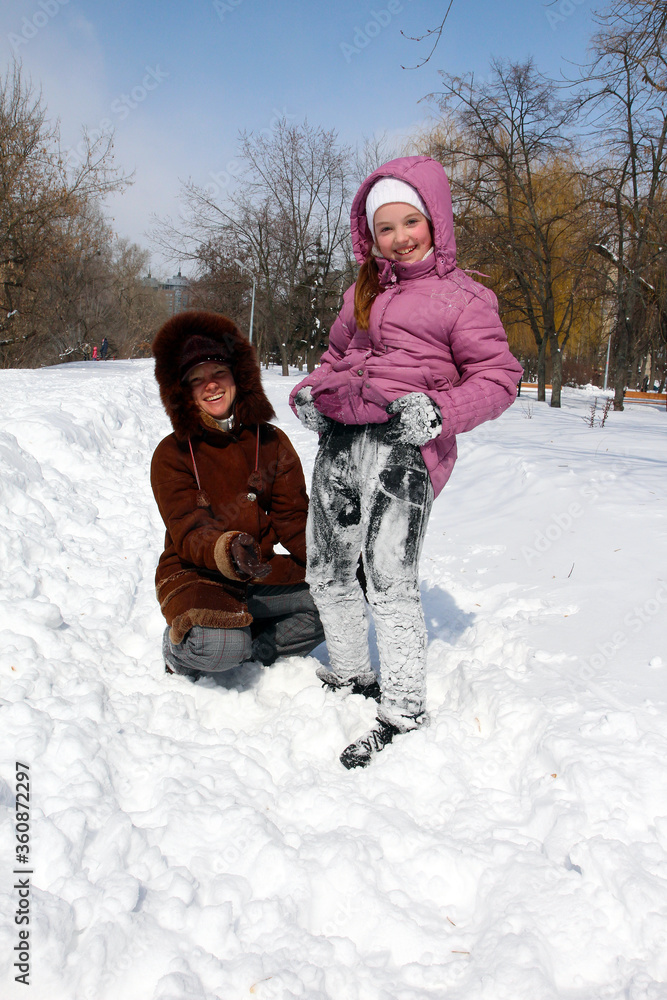 Obraz premium a small smiling girl in snow-covered pants next to a smiling young woman squatting on the snow on a frosty day