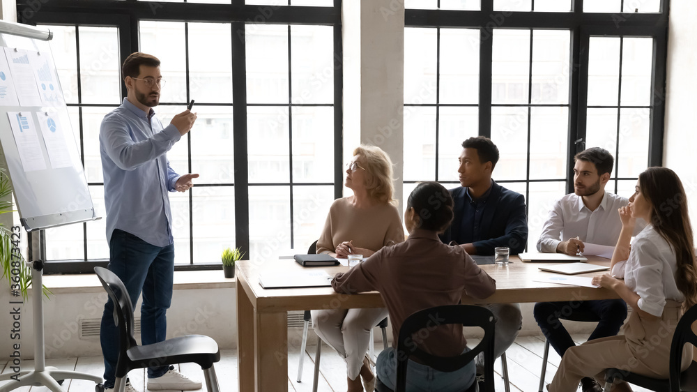 Confident businessman wearing glasses leading corporate meeting with diverse staff, making flip chart presentation, business coach mentor training employees, explaining strategy, presenting stats Confident businessman wearing glasses leading corporate meeting with diverse staff, making flip chart presentation, business coach mentor training employees, explaining strategy, presenting stats