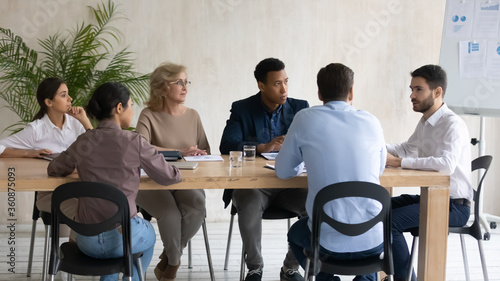 Interested diverse employees team listening to team leader mentor at meeting, sitting at table in modern boardroom, business coach explaining strategy, giving instructions to workers at briefing