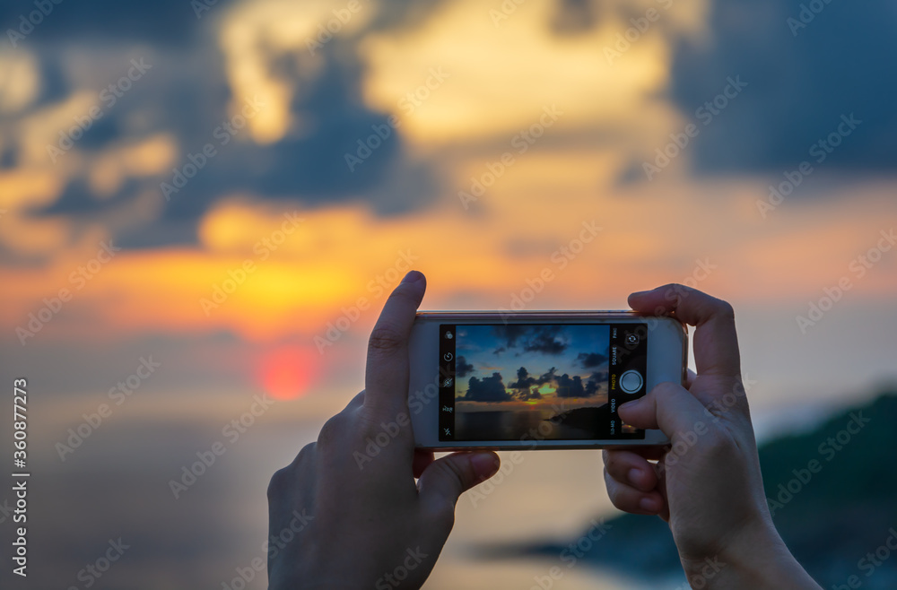 Girl's hand hold a smartphone to take a photography of sunset scenery.