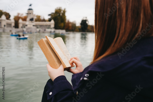 atractiva mujer joven pelirroja con pecas sentada en un banco leyendo y en parque pensativa delante de un estanque © Jake Jakab