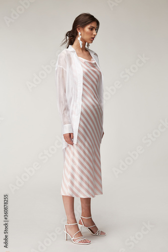 Sexy fashion model beautiful woman pretty face makeup, tanned skin wear summer collection clothes style for romantic date walk trendy light silk in white pink stripes long dress white shirt earrings.