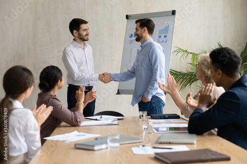 Confident executive businessman wearing glasses shaking successful employee hand, congratulating with job promotion or thanking for work result at corporate meeting, diverse colleagues applauding