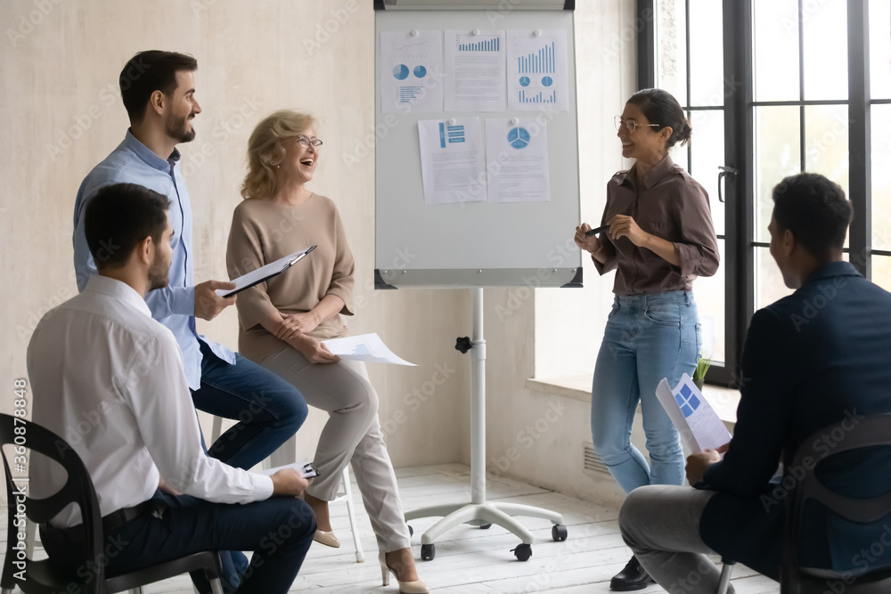 Diverse employees having fun with Indian mentor at corporate meeting, laughing at joke, coach giving flip chart presentation at briefing, presenting financial report, project statistics Diverse employees having fun with Indian mentor at corporate meeting, laughing at joke, coach giving flip chart presentation at briefing, presenting financial report, project statistics