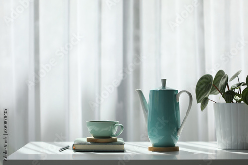 Teal cup and tall teapot with notebook and silver pen on white table with plant pot in front of a see-through curtain 
