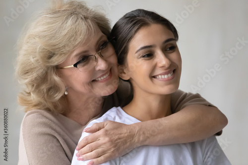 Close up head shot happy middle aged mother wearing glasses hugging adult daughter from back, expressing love and care, smiling positive mature grandmother and granddaughter enjoying tender moment