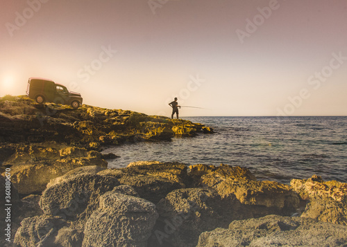 Travelling by car. Young man fishes in the sea from a stone shore. Warm soft colors, romantic style.