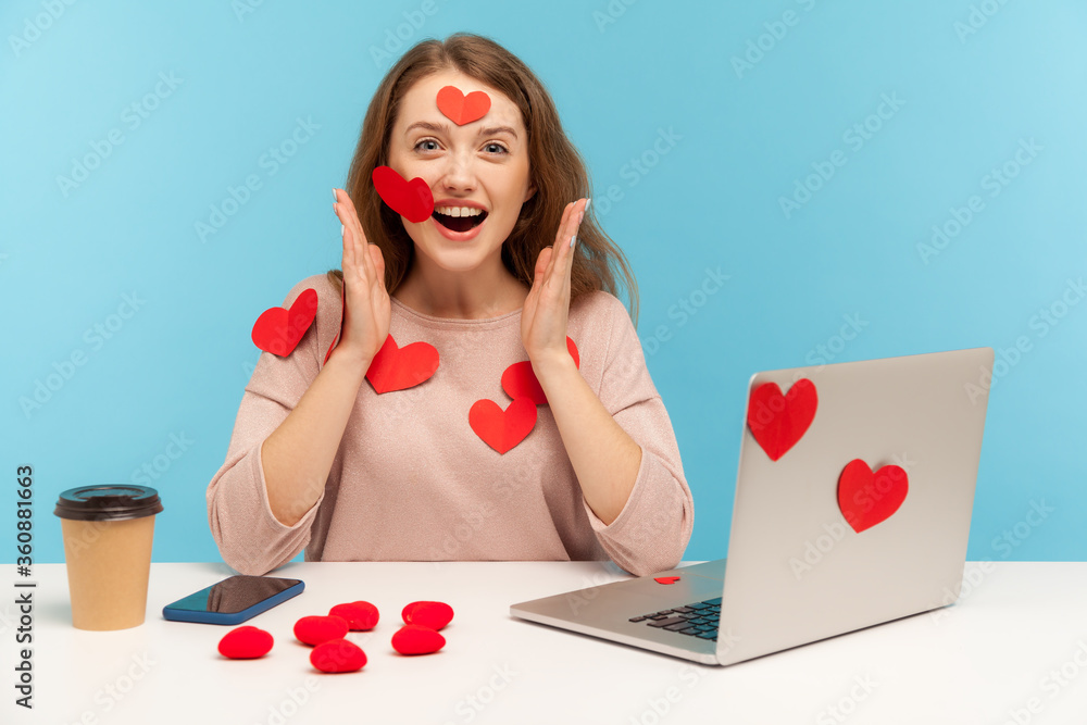 Amazed joyful young woman sitting all covered with sticker love hearts, looking at camera with surprised expression, full of valentine's day greetings. indoor studio shot isolated on blue background Amazed joyful young woman sitting all covered with sticker love hearts, looking at camera with surprised expression, full of valentine's day greetings. indoor studio shot isolated on blue background