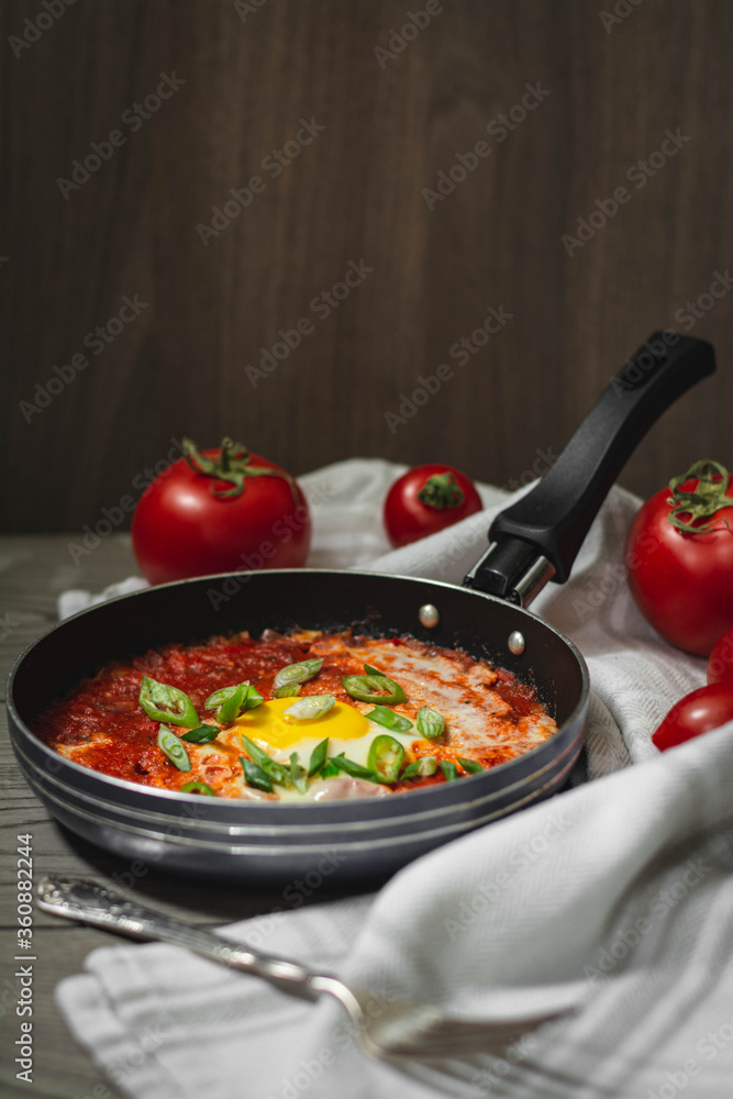 Shakshuka traditional middle eastern dish with tomatoes and eggs in a small pan garnished with scallions and chilli. Tomatoes in the background, dark and moody concept
