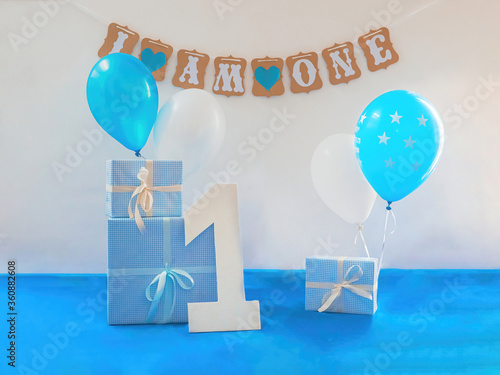 One year birthday decorations set. Decoration for a boy holiday party. Bunch of blue & white balloons, gift boxes, large white kappa board number one shape, and sign with the inscription I am one
