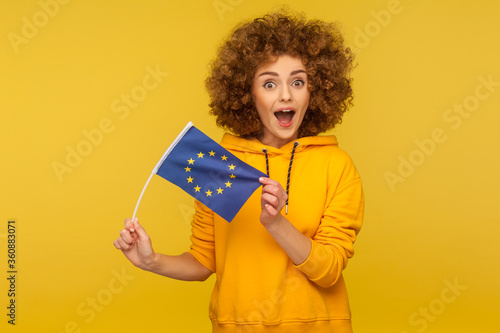 Immigration and citizenship in European Union. Portrait of surprised curly-haired woman in urban style hoodie showing EU flag and looking with amazement. studio shot isolated on yellow background