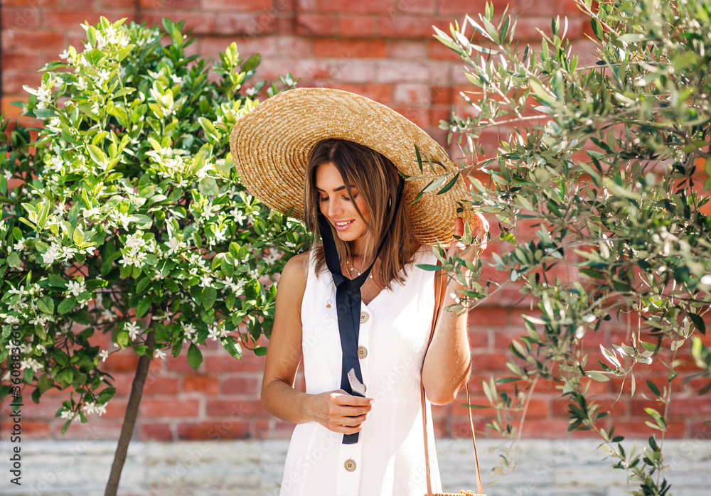 Stylish woman with straw hat standing between two trees and looking down