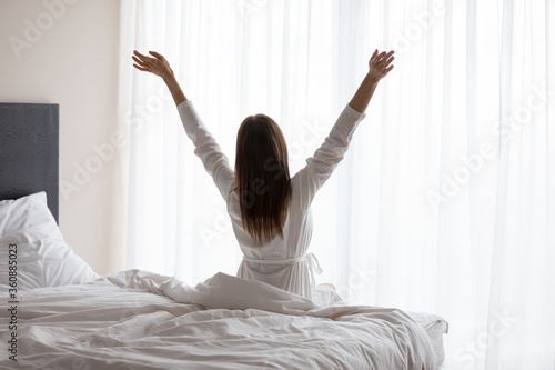 Back view of happy young woman in white bathrobe sit on cozy bed wake up in the morning welcome new sunny day, excited female awaken in bright hotel or home bedroom stretch do morning exercise