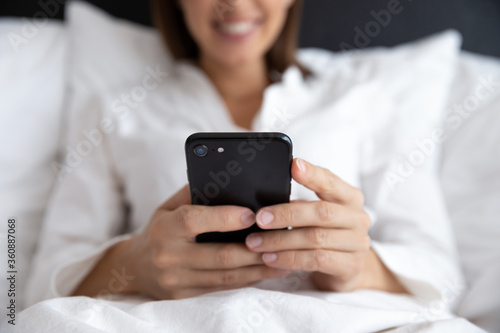 Close up of smiling young woman lying in comfortable bed wake up in the morning using modern smartphone gadget, happy female awaken in cozy home or hotel bedroom browse Internet on cell