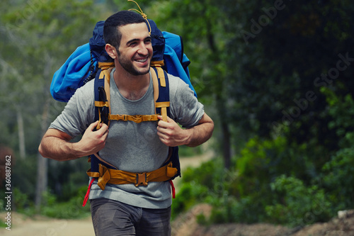 Portrait of a young smiling wanderer man with backpack goes on up the road with copy space, happy hiker walking lonely in mountains at summer day, traveler enjoying his recreation time in nature