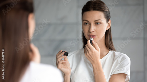 Beautiful millennial woman look in mirror in bathroom do morning every day make up, pretty young female paint color lips with nude lipstick, beauty routine procedures, cosmetics concept