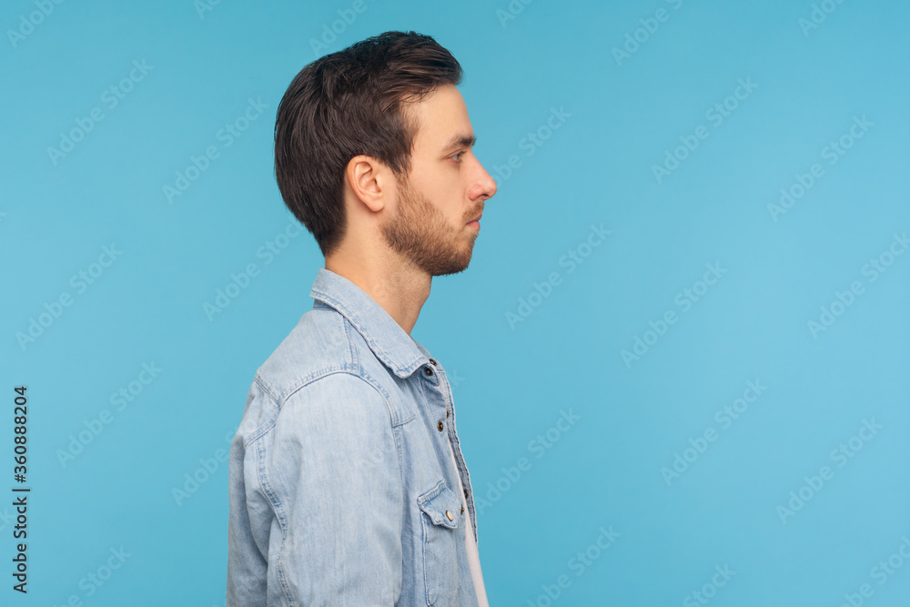Side view of serious handsome young man with stylish hairdo and bristle, wearing worker denim shirt, standing with calm attentive focused expression. indoor studio shot isolated on blue background Side view of serious handsome young man with stylish hairdo and bristle, wearing worker denim shirt, standing with calm attentive focused expression. indoor studio shot isolated on blue background