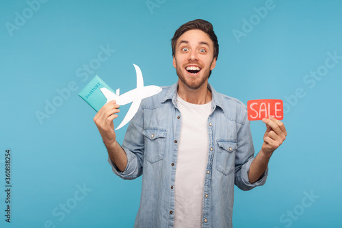 Portrait of amazed happy tourist man in denim shirt holding paper plane, passport and Sale word, planning vacation abroad with low cost air transportation. studio shot isolated on blue background