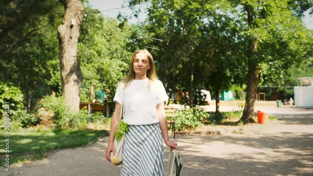 Young woman in casual outfit with food in mesh organic shopping bags is walking in city park. Seeing someone, smiling and waving hand. Slow motion Young woman in casual outfit with food in mesh organic shopping bags is walking in city park. Seeing someone, smiling and waving hand. Slow motion