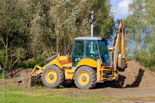 Parked large yellow multifunctional wheel tractor on the glade against green trees in sunny day