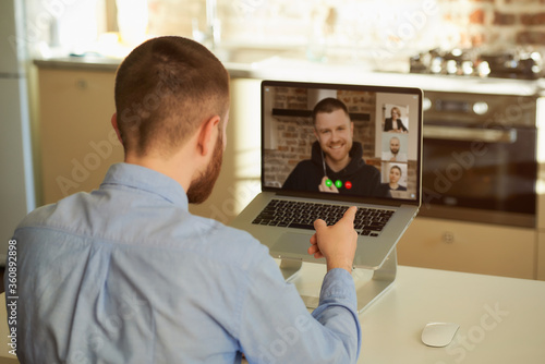 Back view of a boss in a blue shirt who works remotely listening to his employee about business in a video conference on a laptop computer at home. A multiethnic business team at an online meeting.