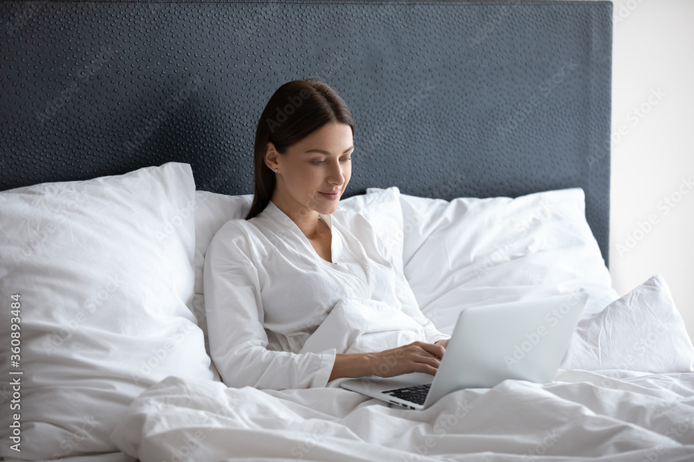Concentrated young woman wake up rest in white bed working typing on laptop, focused millennial female awaken relax in hotel or home bedroom using browsing modern computer in the morning Concentrated young woman wake up rest in white bed working typing on laptop, focused millennial female awaken relax in hotel or home bedroom using browsing modern computer in the morning