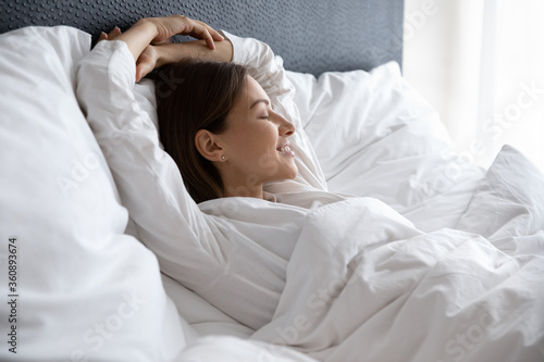 Happy millennial female lying resting in comfortable white bed see dreams napping in the morning, smiling young woman relax sleep on soft pillow in cozy home or hotel bedroom, relaxation concept