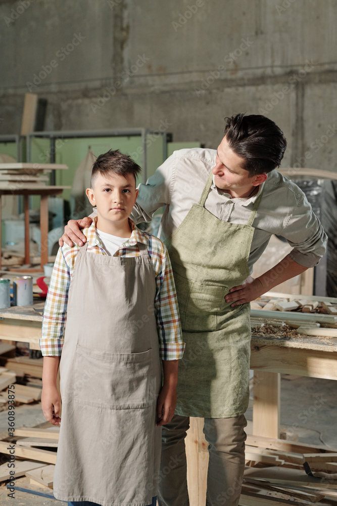 Middle-aged carpenter in apron touching shoulder of son while supporting him in workshop