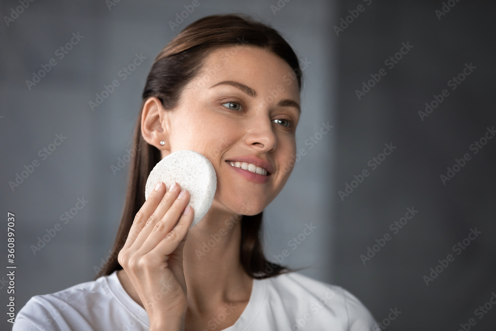 Smiling millennial female look in the mirror massage healthy glowing skin with sponge, happy young woman do daily facial peeling, treatment beauty procedures at home, skincare concept Smiling millennial female look in the mirror massage healthy glowing skin with sponge, happy young woman do daily facial peeling, treatment beauty procedures at home, skincare concept