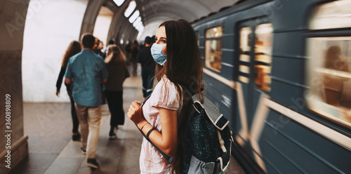 Coronavirus pandemic. Back view of young girl with backpack, wearing protective medical face mask against covid-19. Standing at subway station, waiting for train. Panoramic banner view.