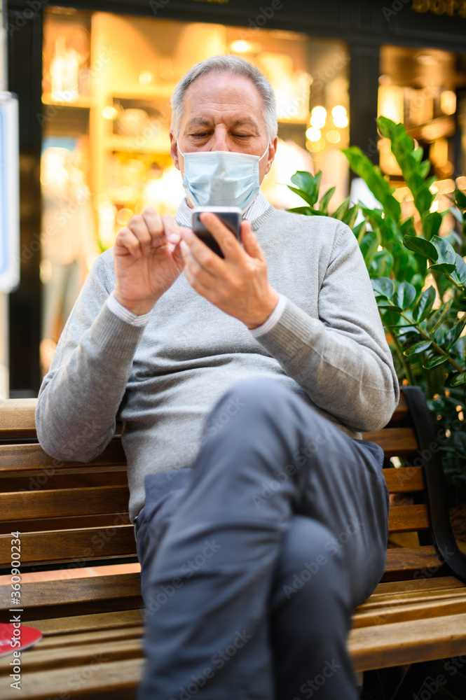Senior man sitting on a bench and using a smartphone in a mall wearing mask, coronavirus concept Senior man sitting on a bench and using a smartphone in a mall wearing mask, coronavirus concept