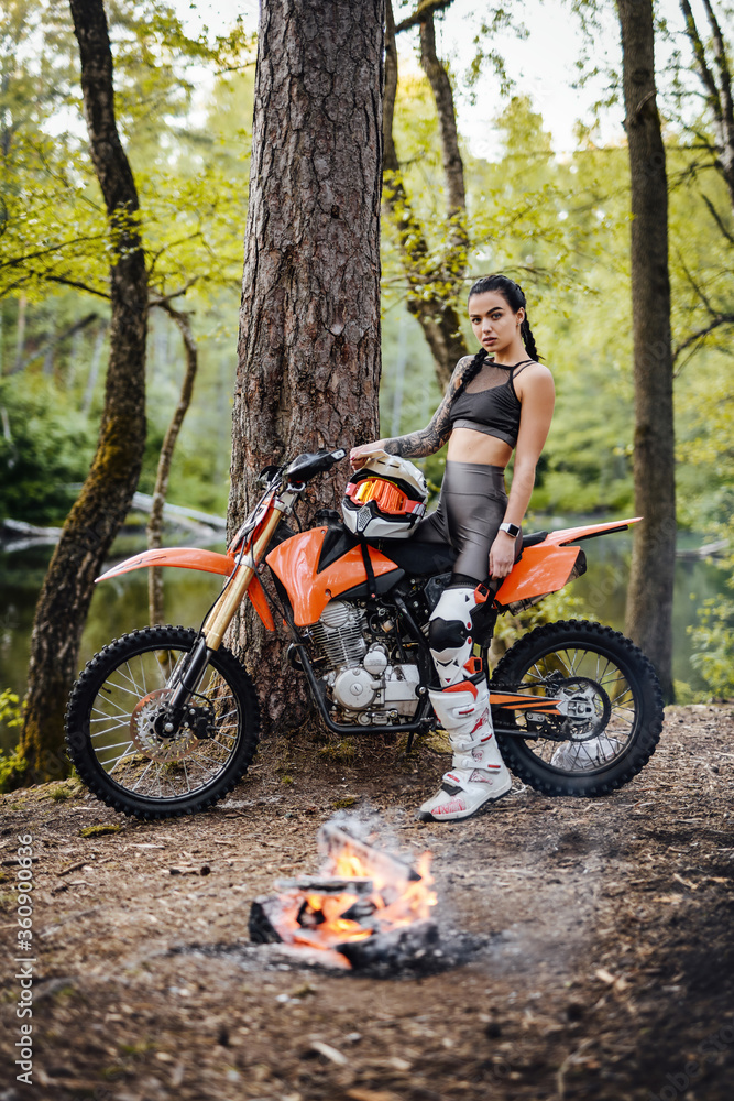 Charming young female racer wearing motocross outfit with semi naked torso sitting on her bike next to the bonfire in the woods