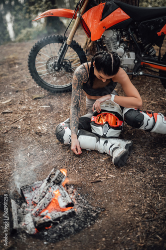 Young tattoed racer girl warming up while sitting next to the bonfire in the woods