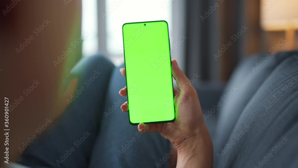 Woman at home lying on a sofa and using smartphone with green mock-up screen in vertical mode. Girl browsing Internet, watching content, videos, blogs. POV. Woman at home lying on a sofa and using smartphone with green mock-up screen in vertical mode. Girl browsing Internet, watching content, videos, blogs. POV.