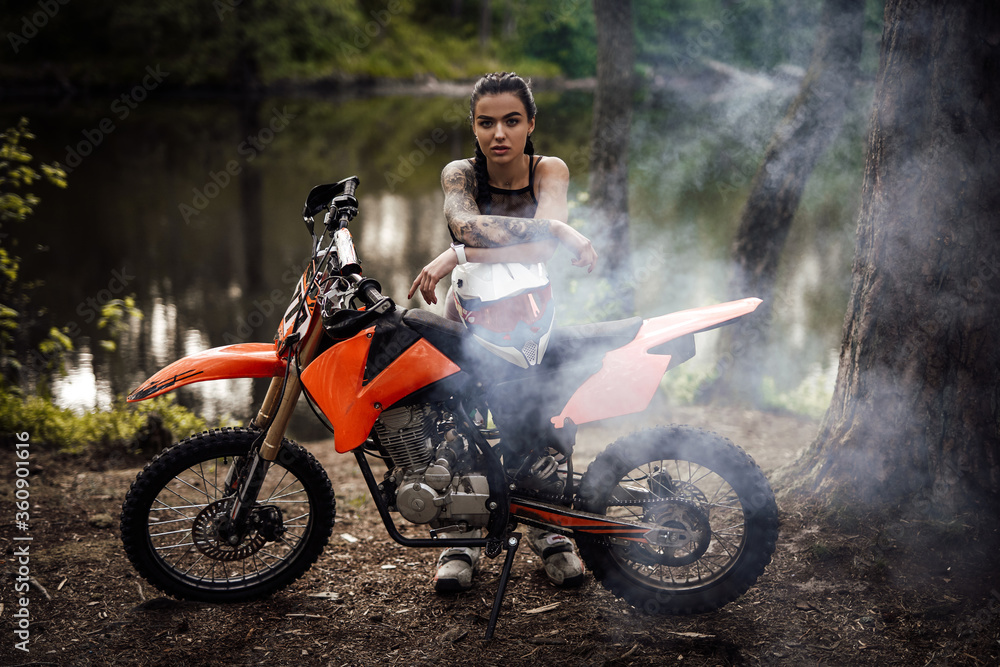 Sensual tattooed racer girl wearing motocross outfit with semi naked torso leaning on her bike and looking on camera in the woods