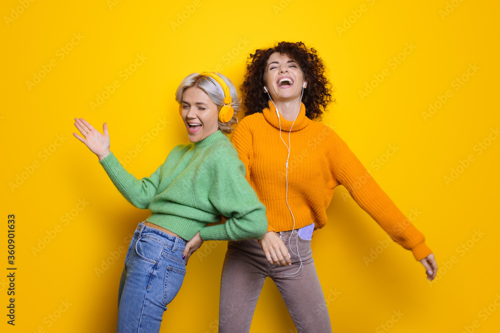 Obraz premium Two happy sisters with curly hair laughing while wearing headphones on a yellow studio wall