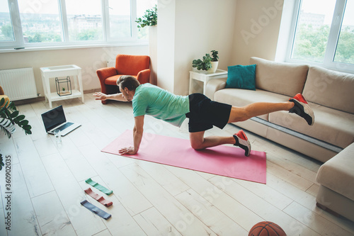 Side view photo of a caucasian man in sportswear stretching on the floor using a laptop and carpet at home
